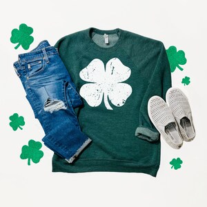 May include: A green sweatshirt with a white four-leaf clover graphic. The sweatshirt is paired with blue jeans and white slip-on shoes. Green shamrocks are scattered around the clothing.