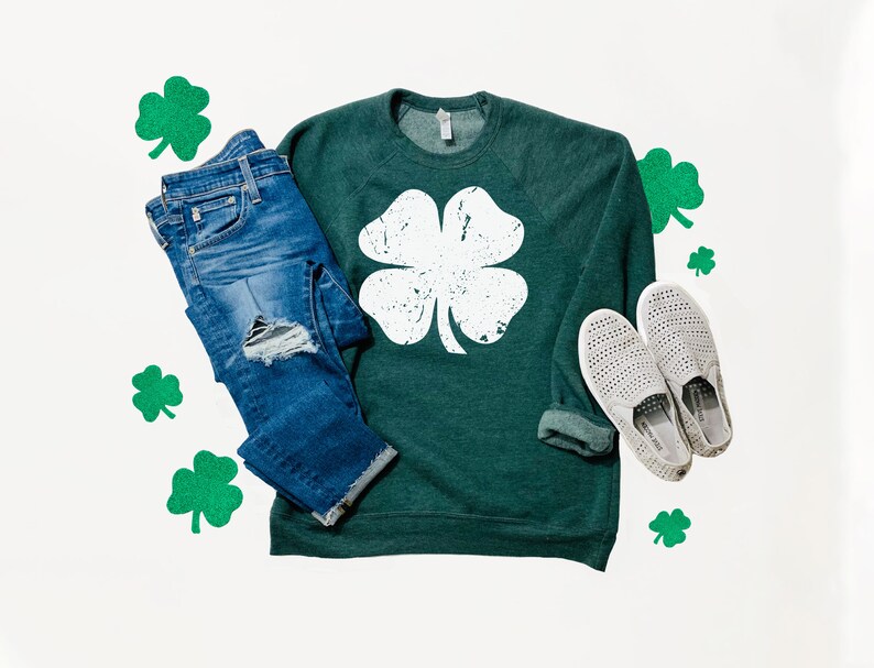 May include: A green sweatshirt with a white four-leaf clover graphic. The sweatshirt is paired with blue jeans and white slip-on shoes. Green shamrocks are scattered around the clothing.