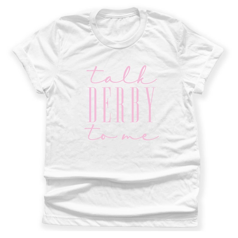 Puede incluir: Camiseta blanca con la frase "talk DERBY to me" en rosa. El texto est&aacute; escrito en cursiva. La camiseta tiene cuello redondo y mangas cortas. El texto est&aacute; centrado.