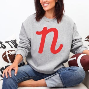 May include: A light gray sweatshirt with a large red letter "n" is the focal point. The person is holding a brown and white football. A decorative pillow with a football design is also visible.