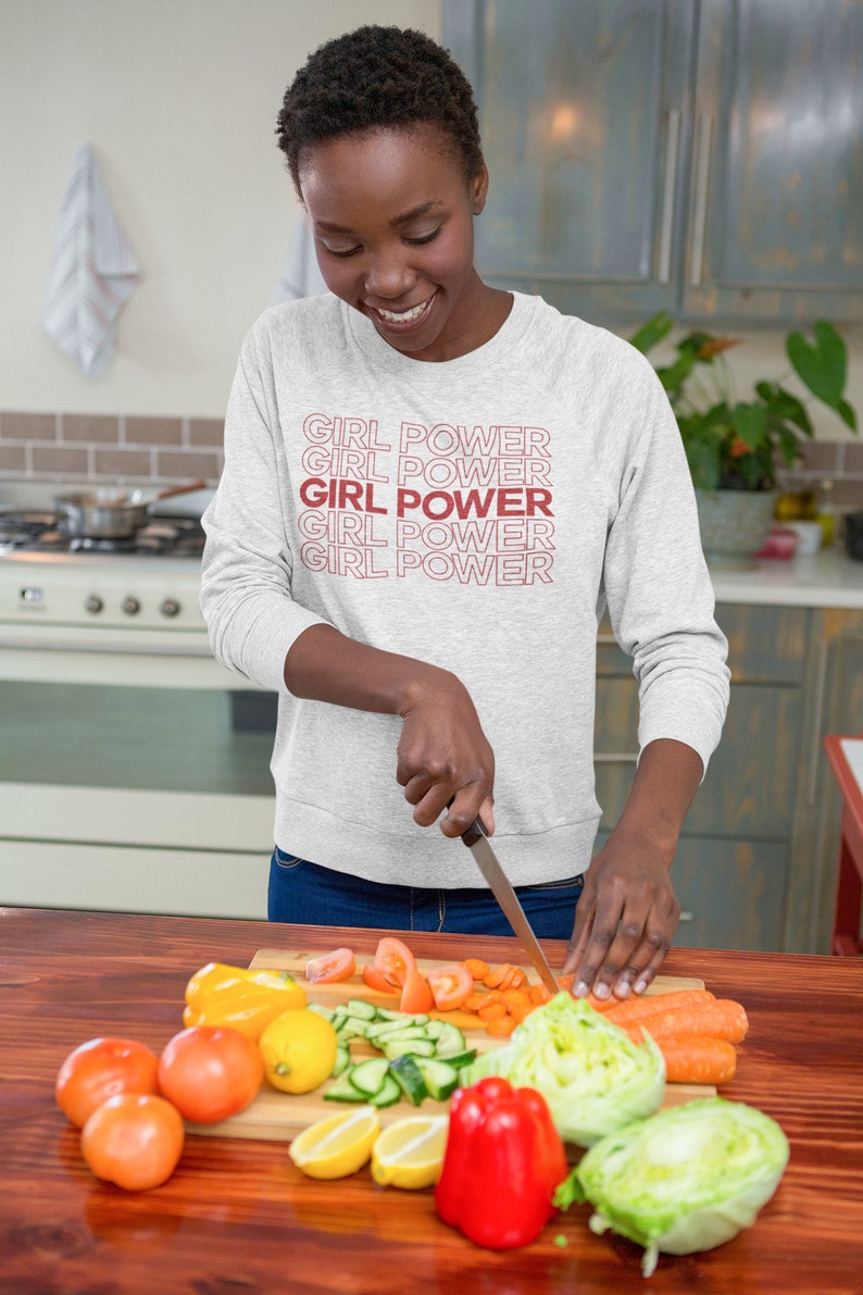 May include: A woman wearing a grey sweatshirt with the text "GIRL POWER" repeated in red. She is chopping vegetables on a wooden cutting board in a kitchen.