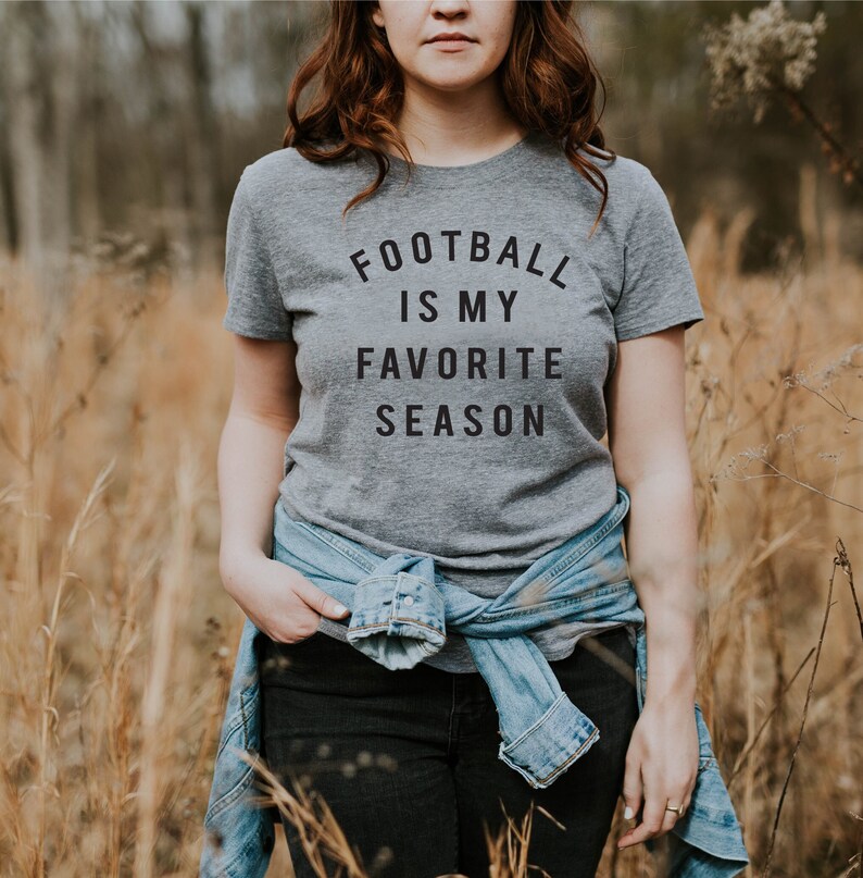 Puede incluir: Una mujer con una camiseta gris con el texto "Football is my favorite season" impreso en ella. Est&aacute; de pie en un campo de hierba alta.