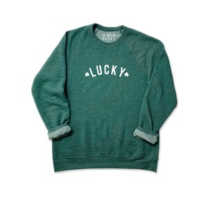 Pu&ograve; includere: Felpa verde con una grafica bianca che dice "LUCKY" con trifogli su entrambi i lati della parola. La felpa ha un collo rotondo e maniche lunghe.