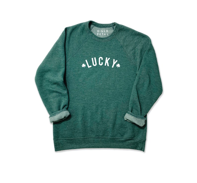 Pu&ograve; includere: Felpa verde con una grafica bianca che dice "LUCKY" con trifogli su entrambi i lati della parola. La felpa ha un collo rotondo e maniche lunghe.