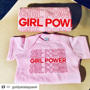 May include: A pink baby bodysuit and matching item, both emblazoned with the red text "GIRL POWER". The bodysuit has the phrase repeated. The accessory also features the phrase. The items are displayed on a light brown surface.