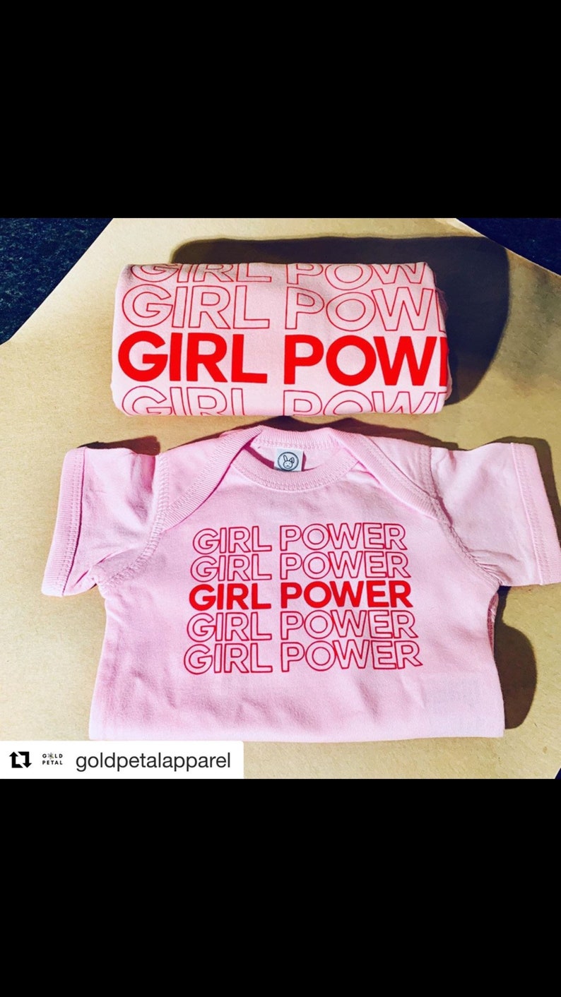May include: A pink baby bodysuit and matching item, both emblazoned with the red text "GIRL POWER". The bodysuit has the phrase repeated. The accessory also features the phrase. The items are displayed on a light brown surface.