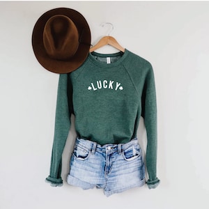 Pu&ograve; includere: Una felpa verde con la scritta "LUCKY" in lettere bianche e simboli di trifoglio su entrambi i lati. La felpa &egrave; appesa a un appendiabiti di legno ed &egrave; abbinata a pantaloncini di jeans.