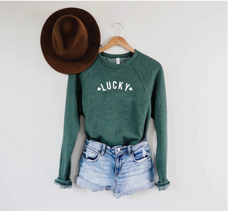 Pu&ograve; includere: Una felpa verde con la scritta "LUCKY" in lettere bianche e simboli di trifoglio su entrambi i lati. La felpa &egrave; appesa a un appendiabiti di legno ed &egrave; abbinata a pantaloncini di jeans.