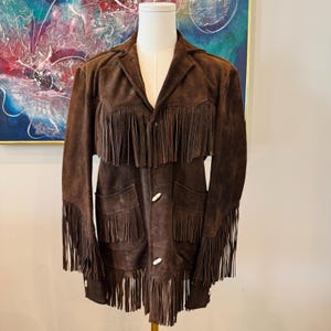 May include: A brown suede jacket with long fringe detailing on the sleeves, chest, and hem. The jacket features two front pockets and three cream-colored buttons. The jacket is displayed on a mannequin.