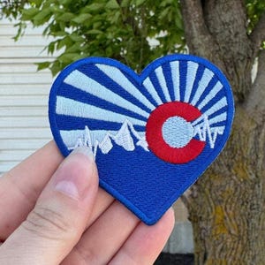 May include: A blue heart-shaped patch featuring a white and blue sunburst design, white mountains, and a red and white circular emblem. The patch has a white embroidered outline and a blue background.