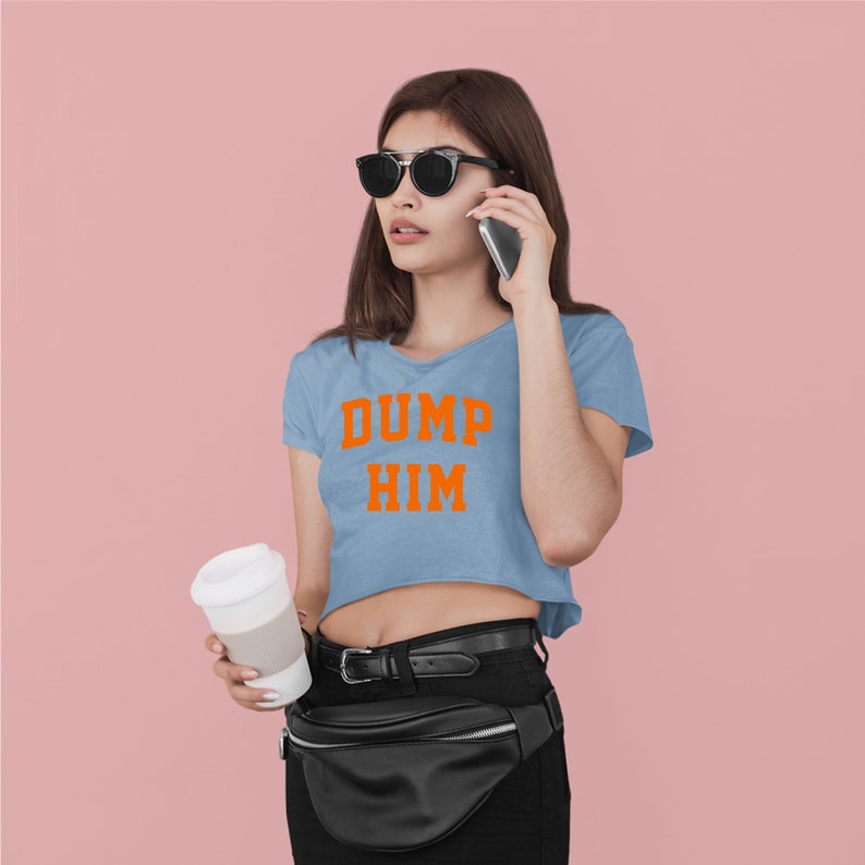 May include: A woman wearing a light blue cropped t-shirt with the text "DUMP HIM" in orange letters. She is wearing black sunglasses, a black belt, and a black fanny pack. She is holding a white to-go cup in her left hand and a cell phone to her ear with her right hand.