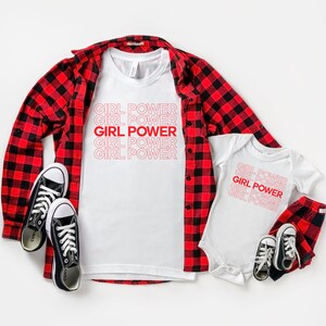 May include: A white t-shirt and baby onesie with the text "GIRL POWER" in red. A red and black plaid shirt and trousers are also present, along with black canvas trainers. The items are arranged on a white surface.