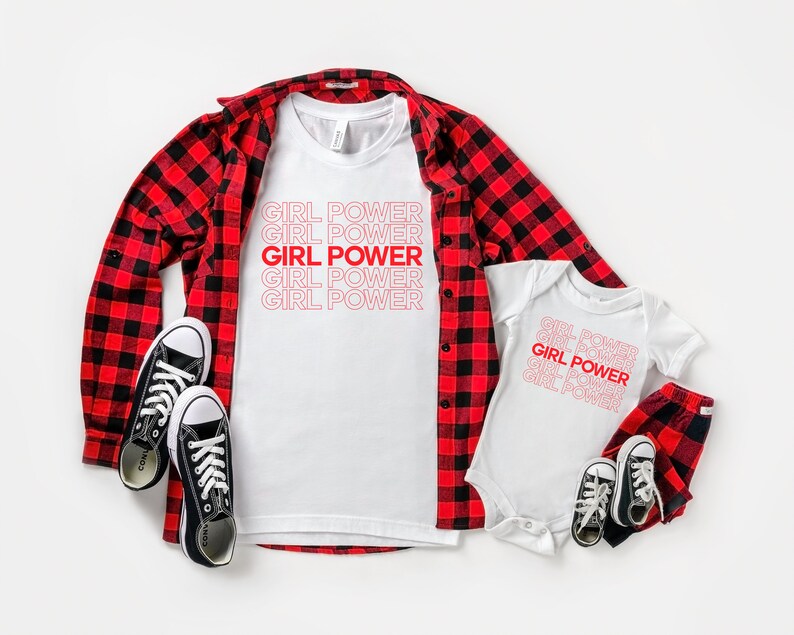 May include: A white t-shirt and baby onesie with the text "GIRL POWER" in red. A red and black plaid shirt and trousers are also present, along with black canvas trainers. The items are arranged on a white surface.
