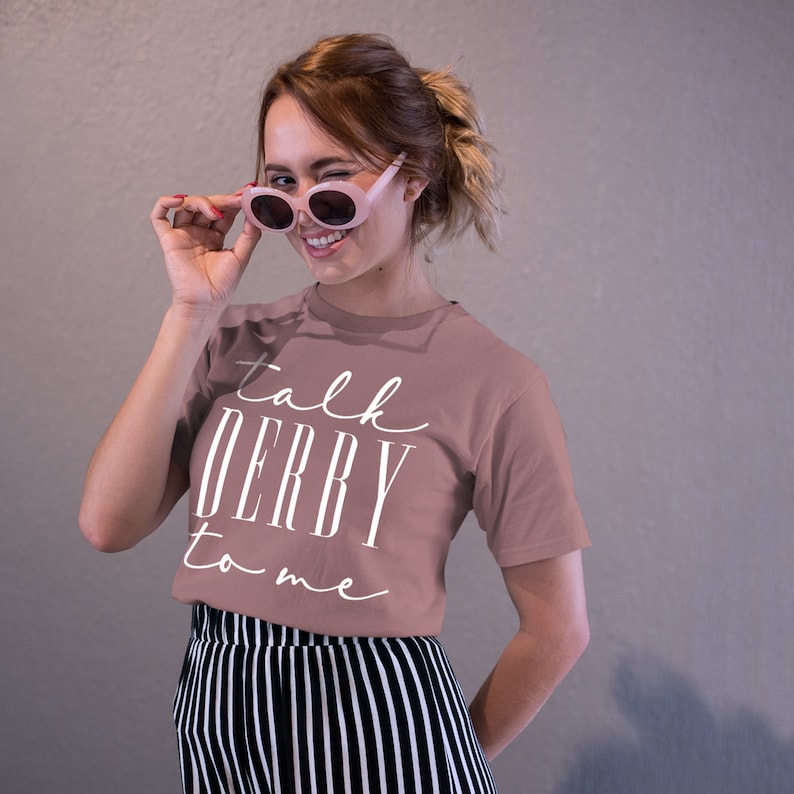 Puede incluir: Una camiseta de color rosa polvoriento con las palabras "talk DERBY to me" en escritura blanca. La modelo lleva gafas de sol rosas y pantalones a rayas blancas y negras.