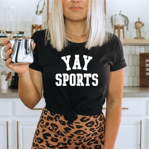 May include: A black t-shirt with white text that reads "YAY SPORTS". The person wearing the shirt is holding a black and white marbled mug.