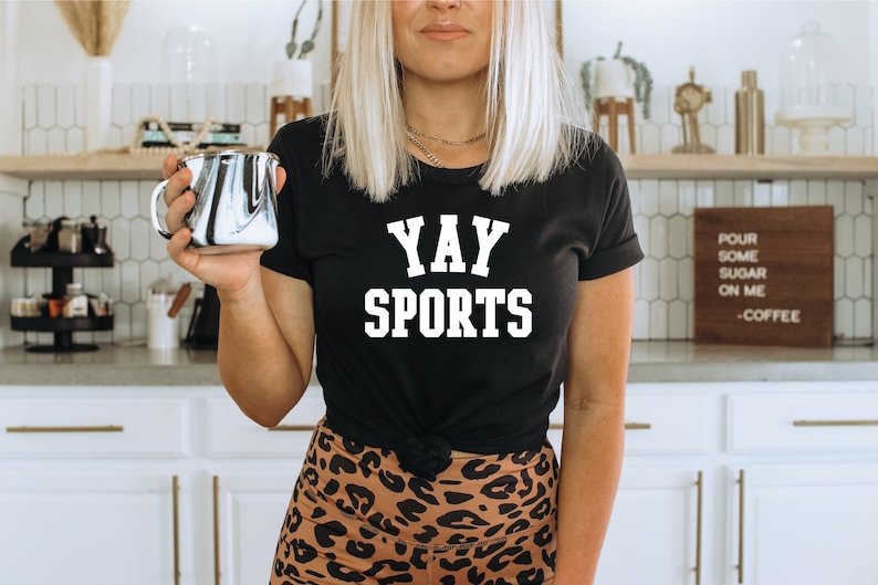 May include: A black t-shirt with white text that reads "YAY SPORTS". The person wearing the shirt is holding a black and white marbled mug.