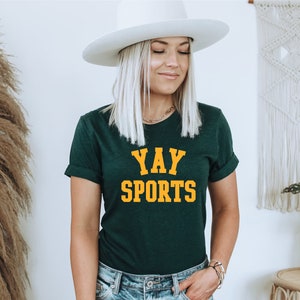 May include: A woman wearing a dark green t-shirt with the text "YAY SPORTS" in yellow letters. She is wearing a white hat and blue jeans.