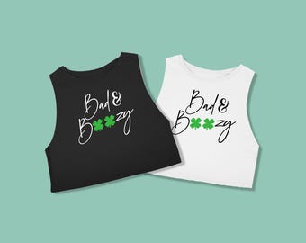 May include: Two sleeveless crop tops, one black and one white, with the phrase "Bad & Boozy" in cursive script and three green shamrocks. The tops are displayed on a light teal background.