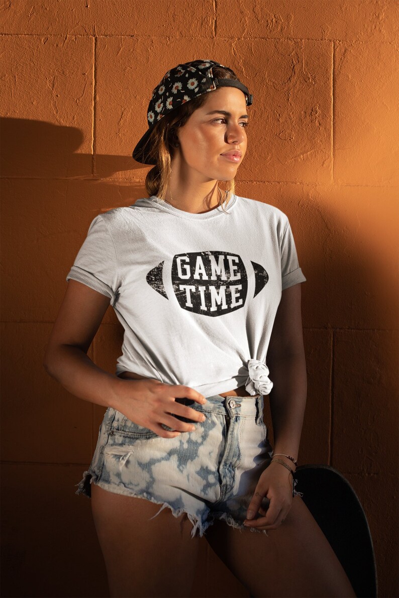 Puede incluir: Una mujer con una camiseta blanca con el texto "GAME TIME" impreso en negro con una fuente desgastada dentro de una forma de bal&oacute;n de f&uacute;tbol. Lleva una gorra de b&eacute;isbol con estampado floral y pantalones cortos de mezclilla.