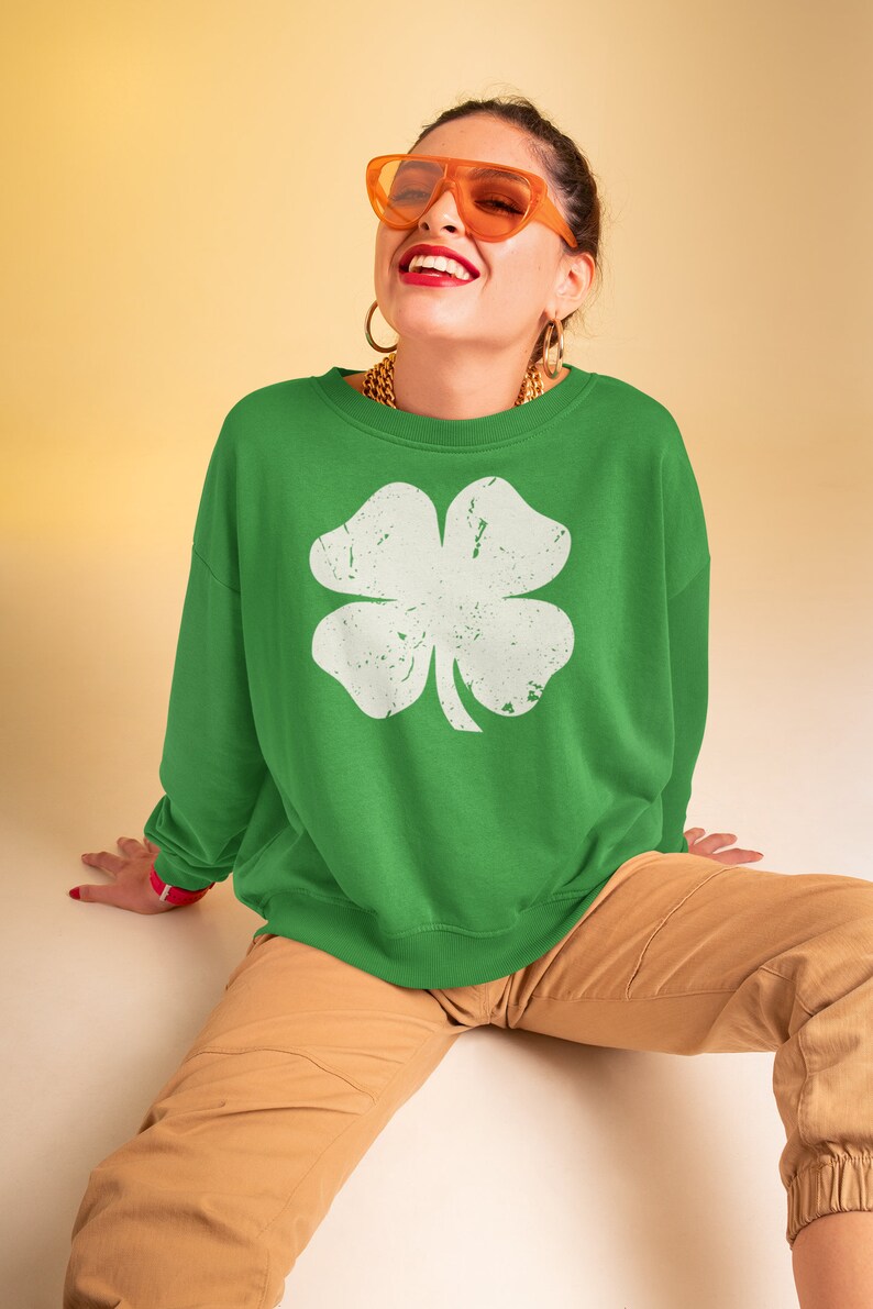 May include: A green sweatshirt with a white four-leaf clover graphic. The model is wearing orange sunglasses and a gold chain necklace.