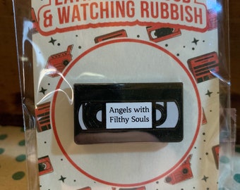 May include: A black enamel pin shaped like a VHS cassette tape with the text "Angels with Filthy Souls" on the label. The pin is on a white card with red text that reads "Eating Junk Food & Watching Rubbish".