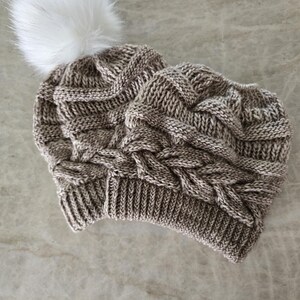 May include: A brown knitted beanie with a white faux fur pom pom on top. The beanie has a cable knit pattern.