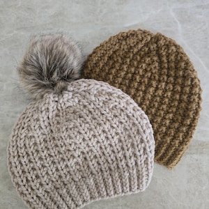 May include: Two knitted hats, one beige with a fluffy pom pom and one brown. Both hats are made with a textured stitch.