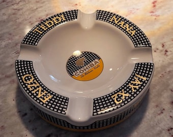 May include: White ceramic ashtray with a black and gold chequered pattern. The ashtray has the words "TEDDI" and "NANA" on the sides and "ENZO" and "IXXI" on the bottom. The centre of the ashtray has a gold logo that says "COHIBA La Habana Cuba".
