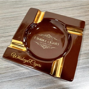 May include: A brown ceramic ashtray with gold trim and the text "I Smoke Cigars That's What I Do And I Know Things" and "Whiskey & Cigars" on the bottom.