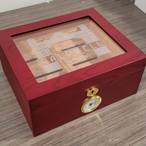 May include: A red wooden cigar humidor with a glass top and a gold-colored hygrometer. The humidor is engraved with the words "CALDWELL" and a building design.