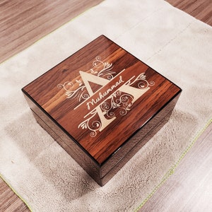 May include: A wooden box with a dark brown finish and a black border. The box has a personalised engraved design with the letter "A" and the name "Muhammad" in a decorative font.