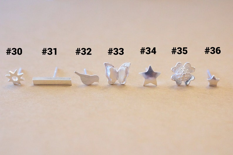May include: A set of 7 silver earrings with different designs, including a sun, a bar, a bird, a butterfly, a star, a flower, and a star. The earrings are numbered 30 through 36.