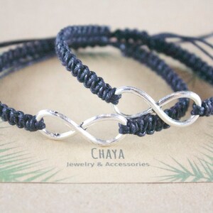 May include: Two dark blue macrame bracelets with silver infinity symbols. The bracelets are adjustable and rest on a card with the text "CHAYA Jewelry & Accessories". The background is a light brown color.