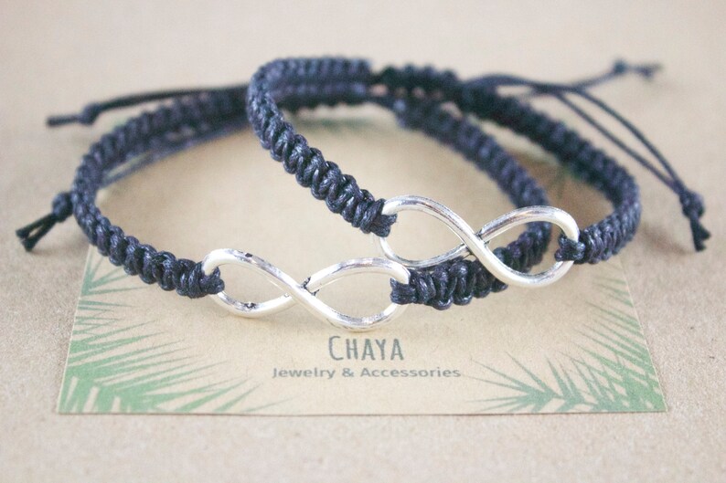 May include: Two dark blue macrame bracelets with silver infinity symbols. The bracelets are adjustable and rest on a card with the text "CHAYA Jewelry & Accessories". The background is a light brown color.