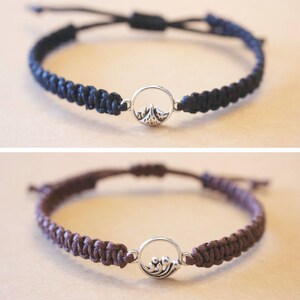 May include: Two braided bracelets with silver charm pendants. The top bracelet is black with a mountain charm. The bottom bracelet is brown with a wave charm.