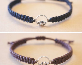 May include: Two braided bracelets with silver charm pendants. The top bracelet is black with a mountain charm. The bottom bracelet is brown with a wave charm.