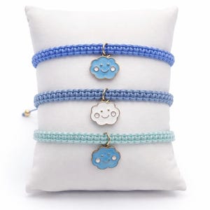 May include: Three macrame bracelets in shades of blue and aqua, each adorned with a smiling cloud charm. The bracelets are displayed on a white pillow, showcasing the different colors and designs. The charms have a gold-colored border.