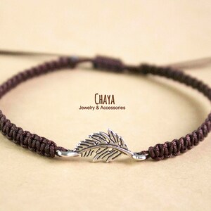 May include: A dark brown macrame bracelet with a silver-colored feather charm. The bracelet is knotted and adjustable. The text "CHAYA Jewelry & Accessories" is visible.