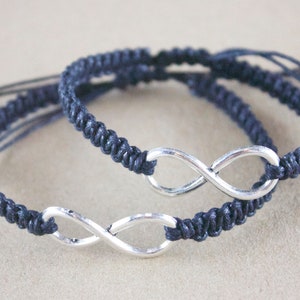 May include: Two navy blue macrame bracelets with silver infinity symbols. The bracelets are adjustable with sliding knots. The background is a neutral tan color. The bracelets are a symbol of everlasting love and friendship.