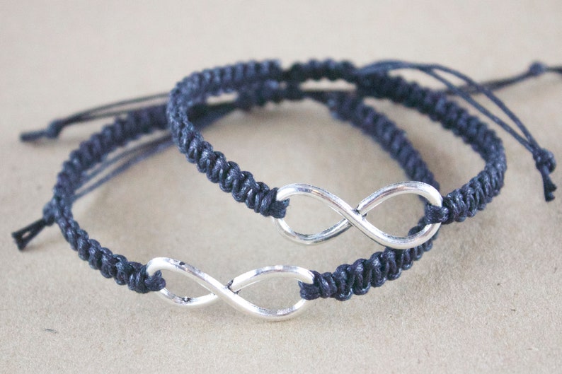 May include: Two navy blue macrame bracelets with silver infinity symbols. The bracelets are adjustable with sliding knots. The background is a neutral tan color. The bracelets are a symbol of everlasting love and friendship.