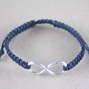 May include: A navy blue macrame bracelet featuring a silver-colored infinity symbol. The bracelet is made of woven cord and has adjustable ties. The infinity symbol is centered on the wristband.