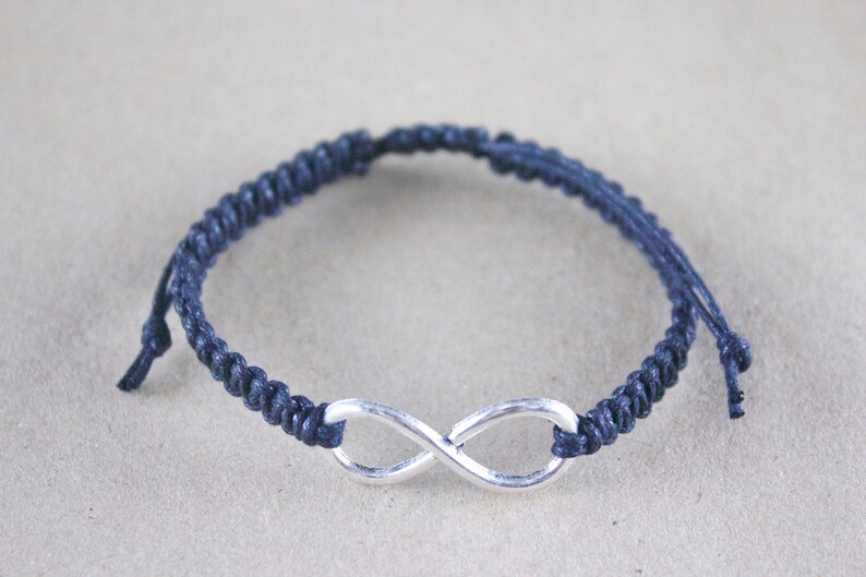 May include: A navy blue macrame bracelet featuring a silver-colored infinity symbol. The bracelet is made of woven cord and has adjustable ties. The infinity symbol is centered on the wristband.