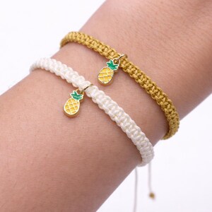 May include: Two handmade bracelets, one cream-colored and one gold, are worn on a wrist. Each bracelet features a small, gold-colored pineapple charm with green and yellow enamel detailing. The bracelets are adjustable.