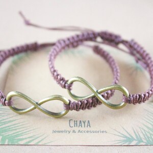 May include: Two matching mauve macrame bracelets with bronze-colored infinity symbols. The bracelets are displayed on a card with the text "CHAYA Jewelry & Accessories". The bracelets are adjustable.