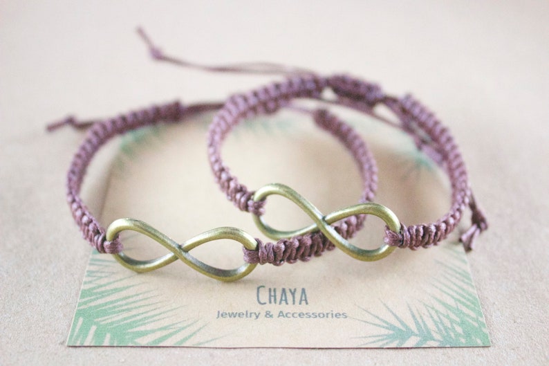 May include: Two matching mauve macrame bracelets with bronze-colored infinity symbols. The bracelets are displayed on a card with the text "CHAYA Jewelry & Accessories". The bracelets are adjustable.