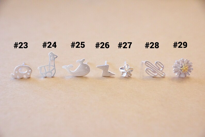 May include: Nine silver earrings with different designs, including an elephant, a giraffe, a whale, a lightning bolt, a flower, a cactus, and a daisy. The earrings are numbered 23 through 29.