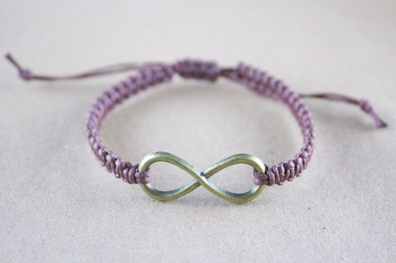 May include: A handmade macrame bracelet in a muted purple color, featuring a bronze-colored infinity symbol. The bracelet is adjustable with sliding knots, offering a comfortable fit. This accessory is perfect for everyday wear.