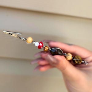 May include: A silver clip with a beaded charm. The beads are brown, white, red, and tiger's eye. The charm is a small red and white mushroom.
