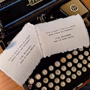 May include: A classic black Erika typewriter with white keys and a paper featuring the quote "I'm so glad I live in a world where there are Octobers." from Anne of Green Gables. The typewriter is on a wooden surface.