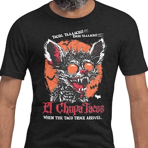 May include: A black t-shirt with a graphic of a cartoon monster with red eyes and fangs. The monster is holding a taco and the text "El Chupa Tacos" is written below the monster. The text "When the taco truck arrives" is written below the monster's name.